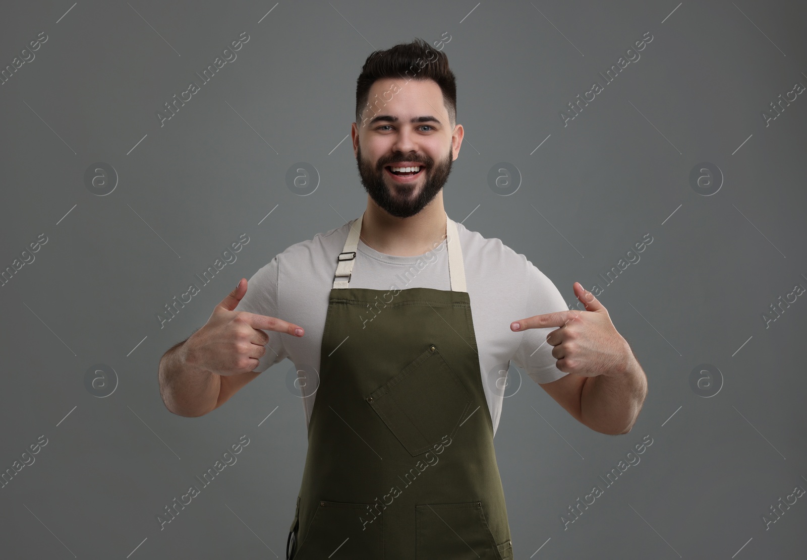 Photo of Smiling man pointing at kitchen apron on grey background. Mockup for design