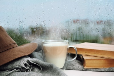 Image of Cup of delicious hot drink, books and scarf on window sill after rain