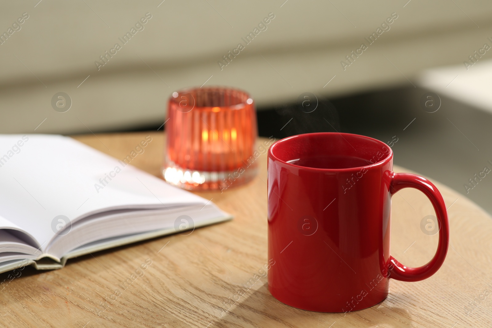 Photo of Red mug with drink, open book and burning candle on wooden table indoors. Mockup for design