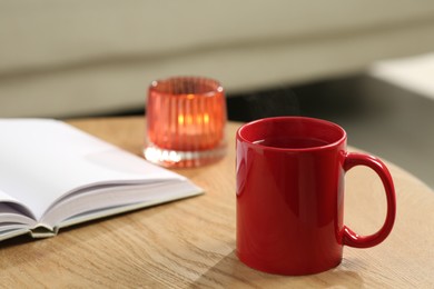 Red mug with drink, open book and burning candle on wooden table indoors. Mockup for design