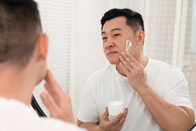 Photo of Handsome man applying cream onto his face near mirror indoors