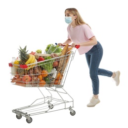 Photo of Young woman in medical mask with shopping cart full of groceries on white background