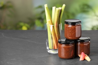 Photo of Jars of tasty rhubarb jam and stalks on grey table, space for text