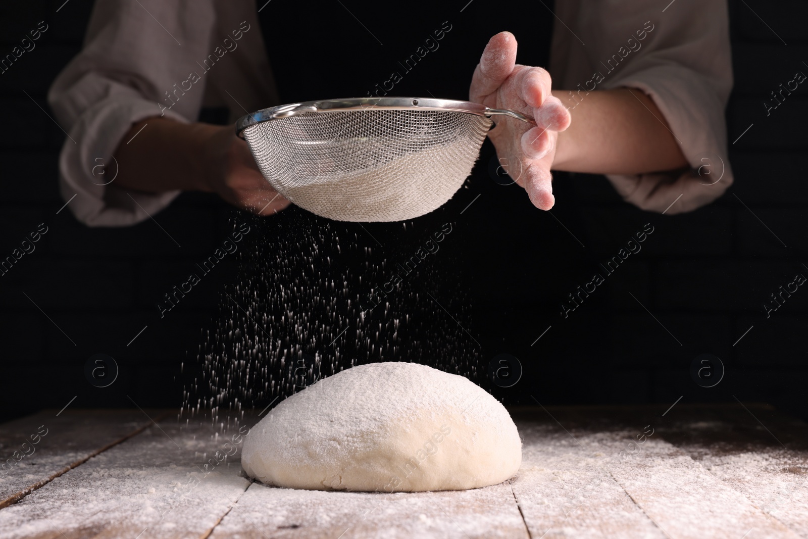 Photo of Woman sprinkling flour over dough at wooden table on dark background, closeup