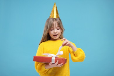 Photo of Cute little girl in party hat opening gift on light blue background