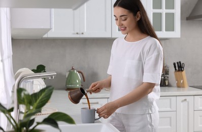 Photo of Beautiful woman pouring coffee from jezve into cup in kitchen. Lazy morning