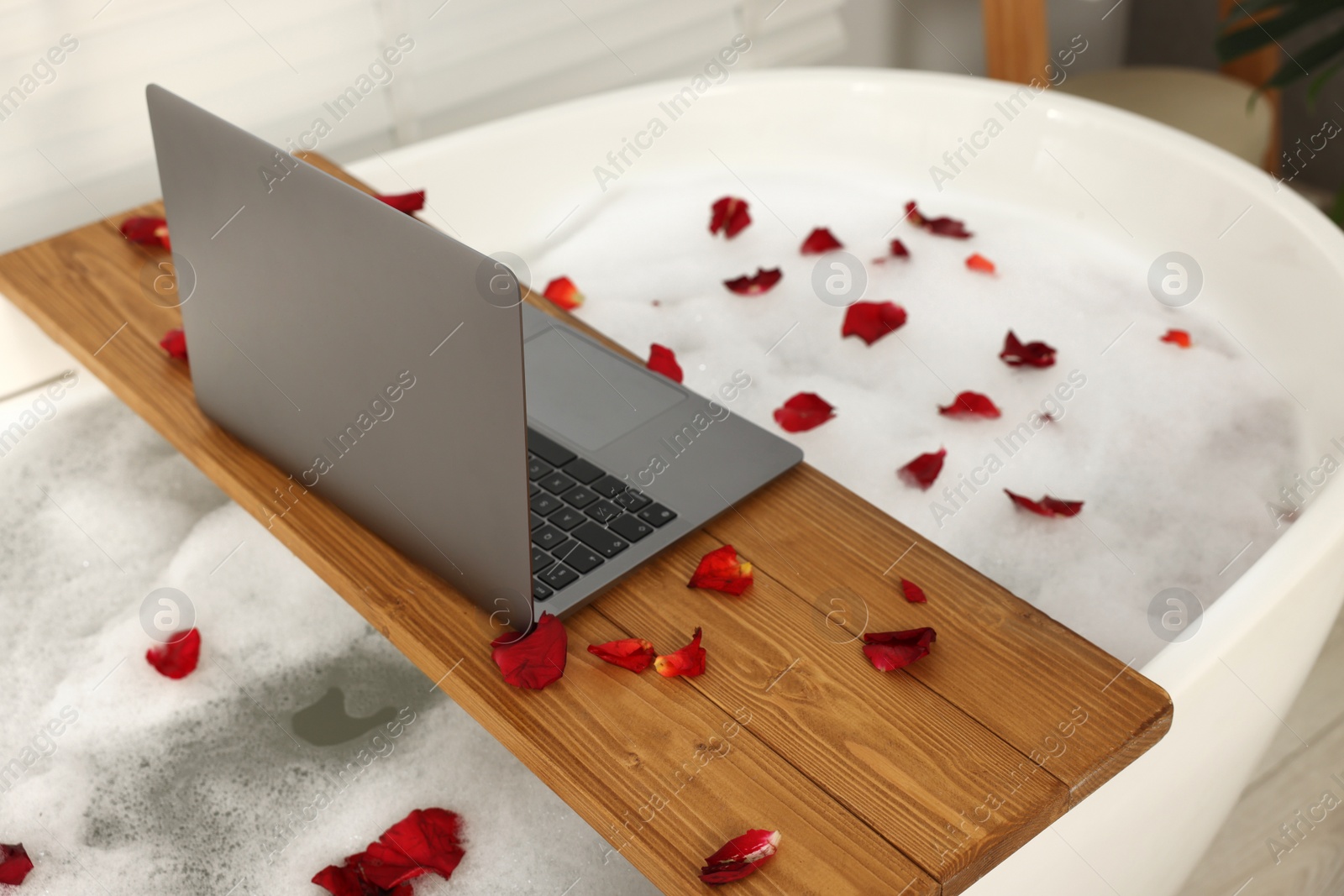 Photo of Wooden board with laptop and rose petals on bath tub