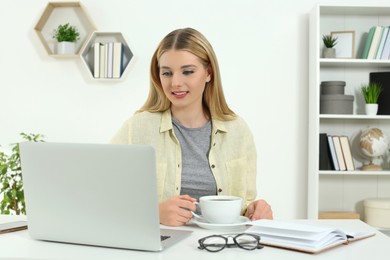 Home workplace. Happy woman with cup of hot drink looking at laptop at white desk in room