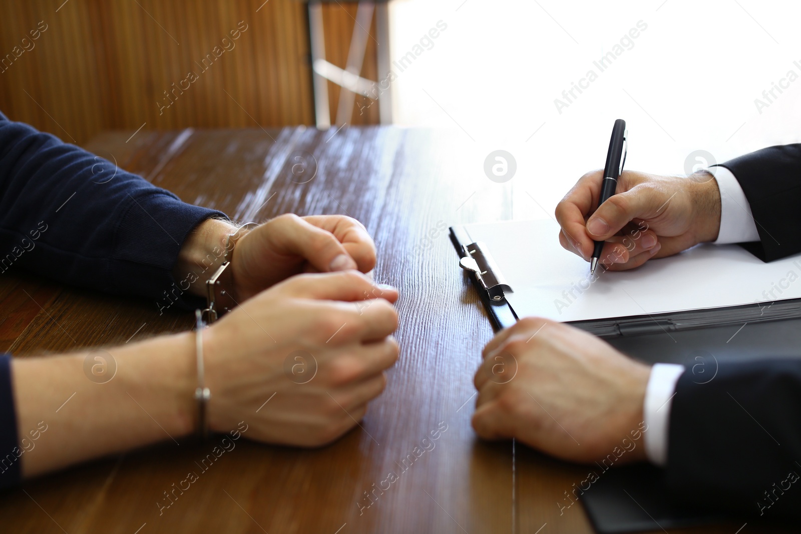 Photo of Police officer interrogating criminal in handcuffs at desk indoors