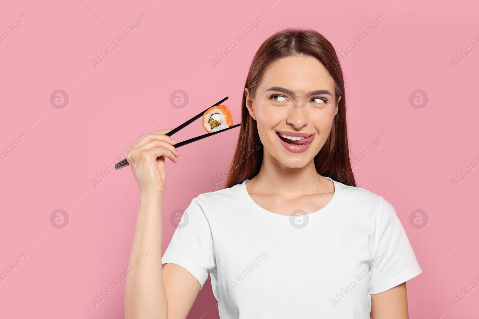 Photo of Beautiful young woman holding sushi roll with chopsticks on pink background