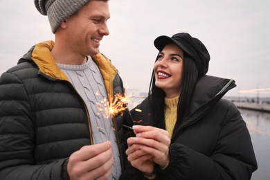 Couple in warm clothes holding burning sparklers near river