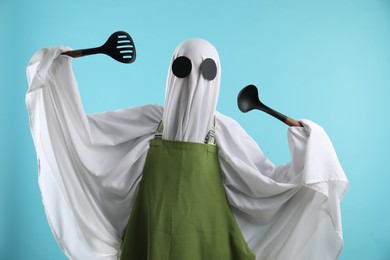 Woman in ghost costume and apron with kitchen set on light blue background