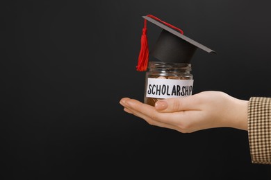 Photo of Woman holding glass jar of coins and graduation cap against black background, closeup with space for text. Scholarship concept