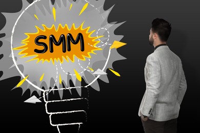 Image of Social media marketing. Man in business attire standing near black wall with illustration of light bulb and abbreviation SMM