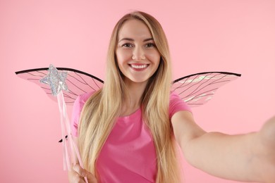 Beautiful girl in fairy costume with wings and magic wand taking selfie on pink background
