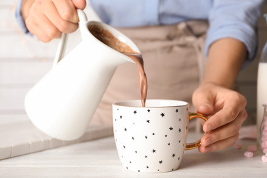 Photo of Woman pouring hot cocoa drink into cup on white wooden table, closeup