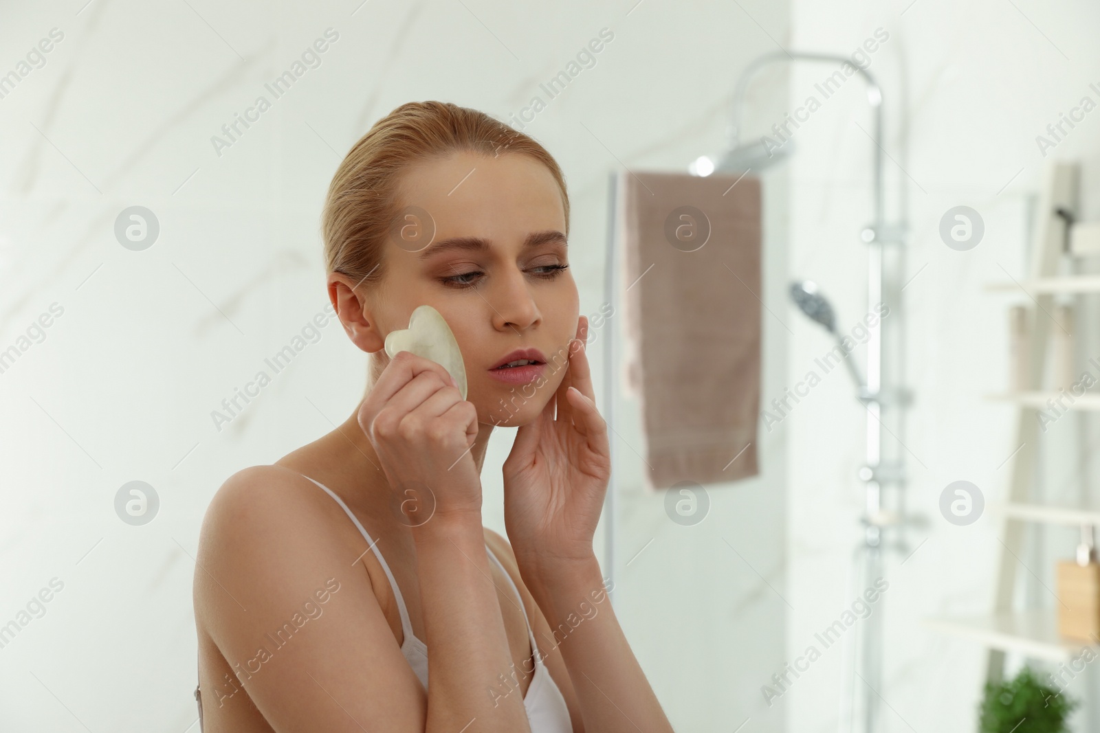 Photo of Beautiful young woman doing facial massage with gua sha tool in bathroom. Space for text
