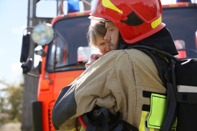 Firefighter in uniform holding rescued little girl near fire truck outdoors. Save life