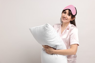 Happy woman in pyjama and sleep mask holding pillow on light grey background, space for text