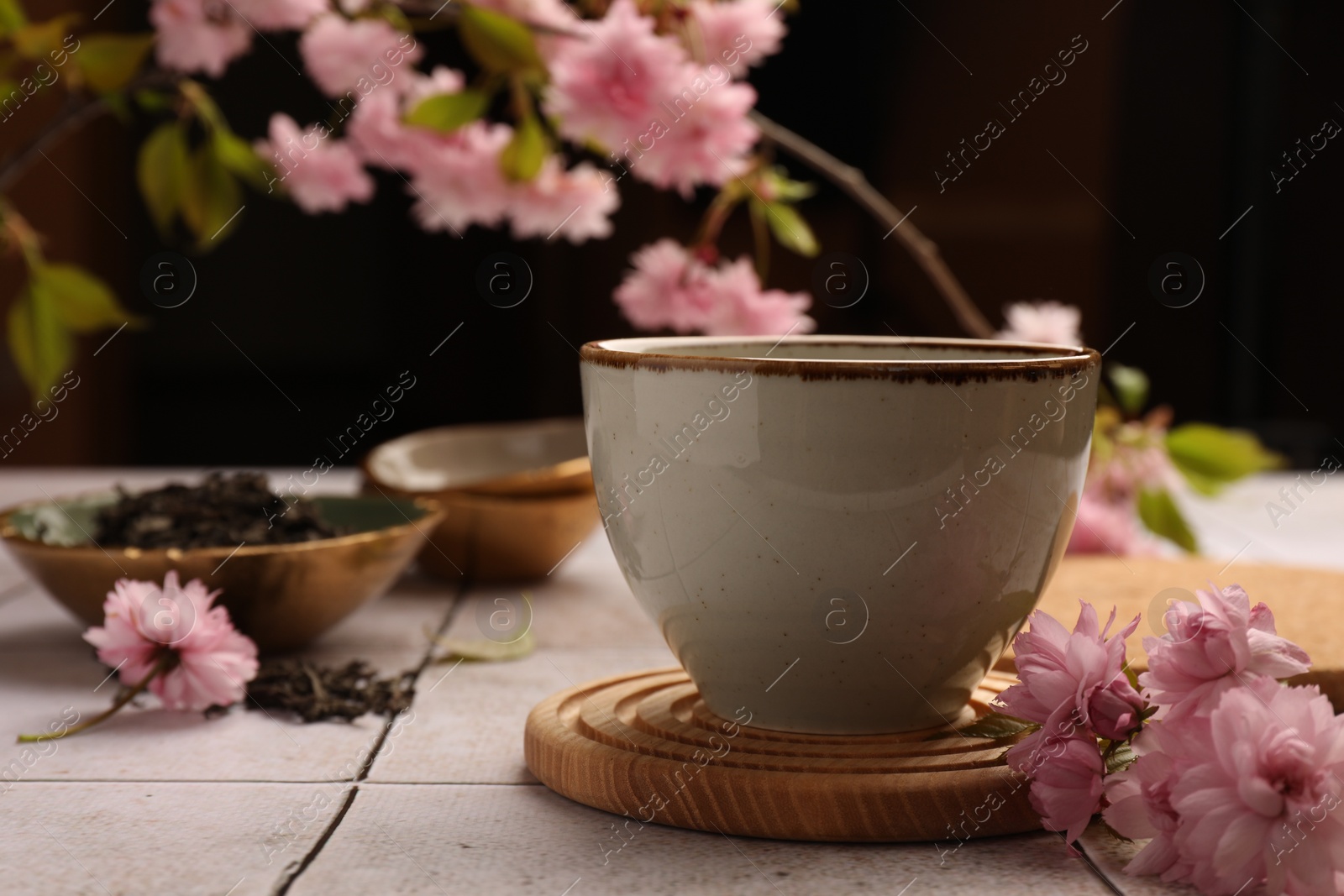 Photo of Traditional ceremony. Cup of brewed tea and sakura flowers on tiled table, closeup