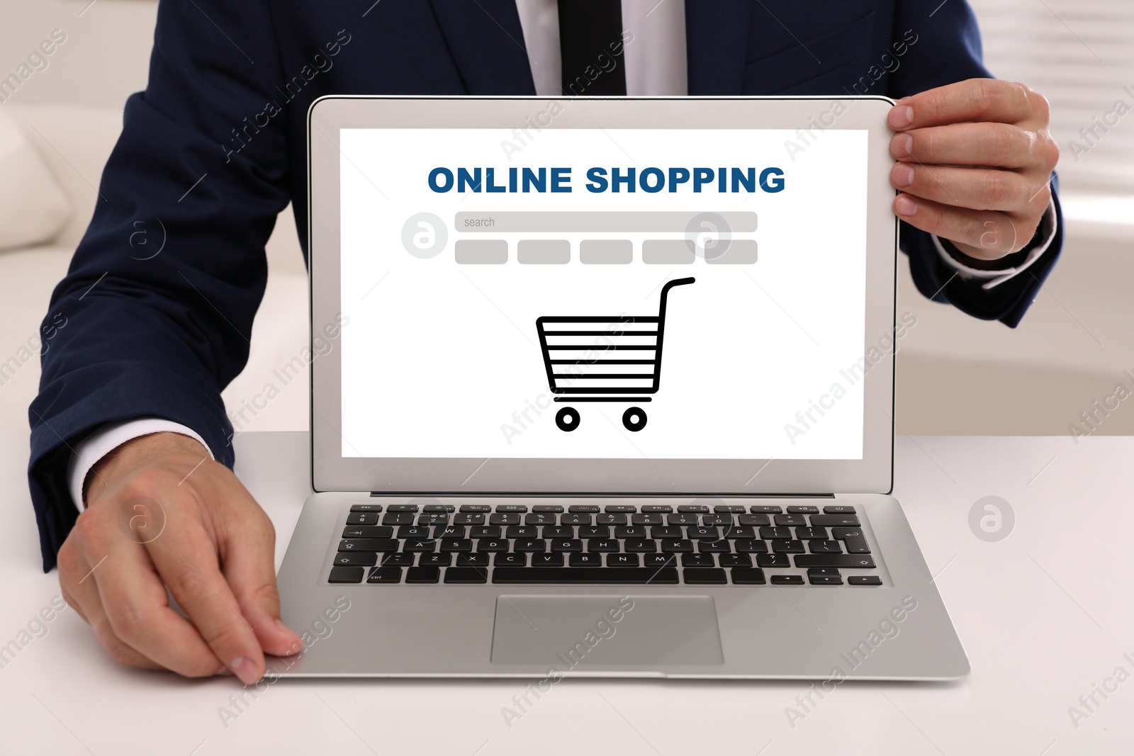 Image of Online shopping. Man showing laptop with search bar and illustration of cart at white table, closeup