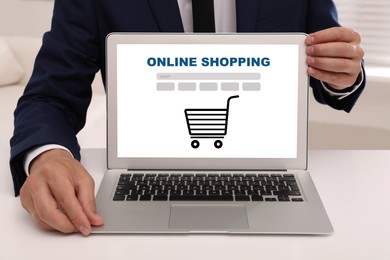 Image of Online shopping. Man showing laptop with search bar and illustration of cart at white table, closeup