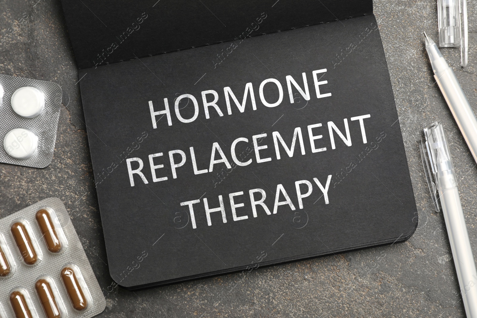 Image of Black notebook with text Hormone Replacement Therapy, pills and pens on grey background, flat lay