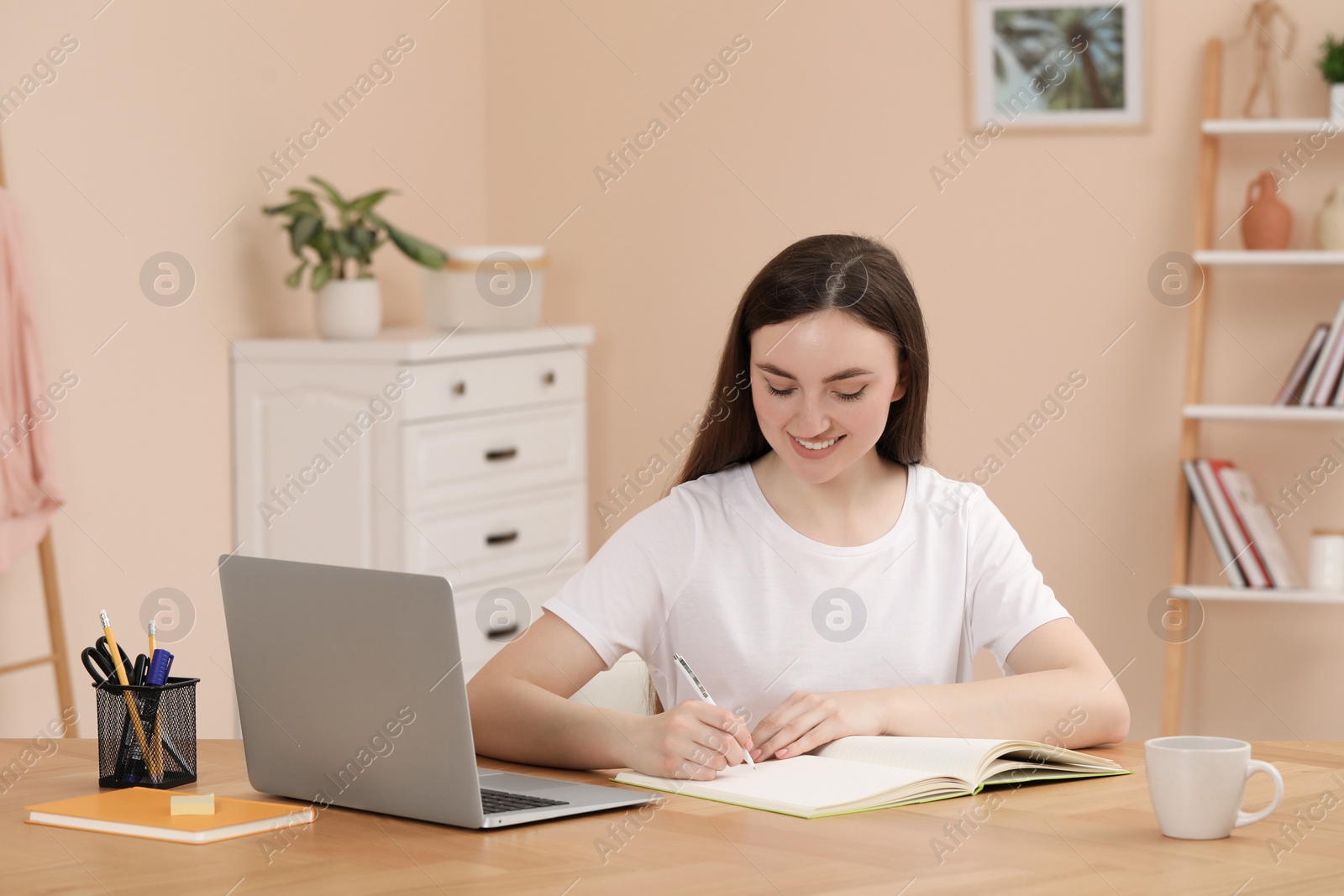 Photo of Woman writing in notebook while working on laptop at wooden table indoors