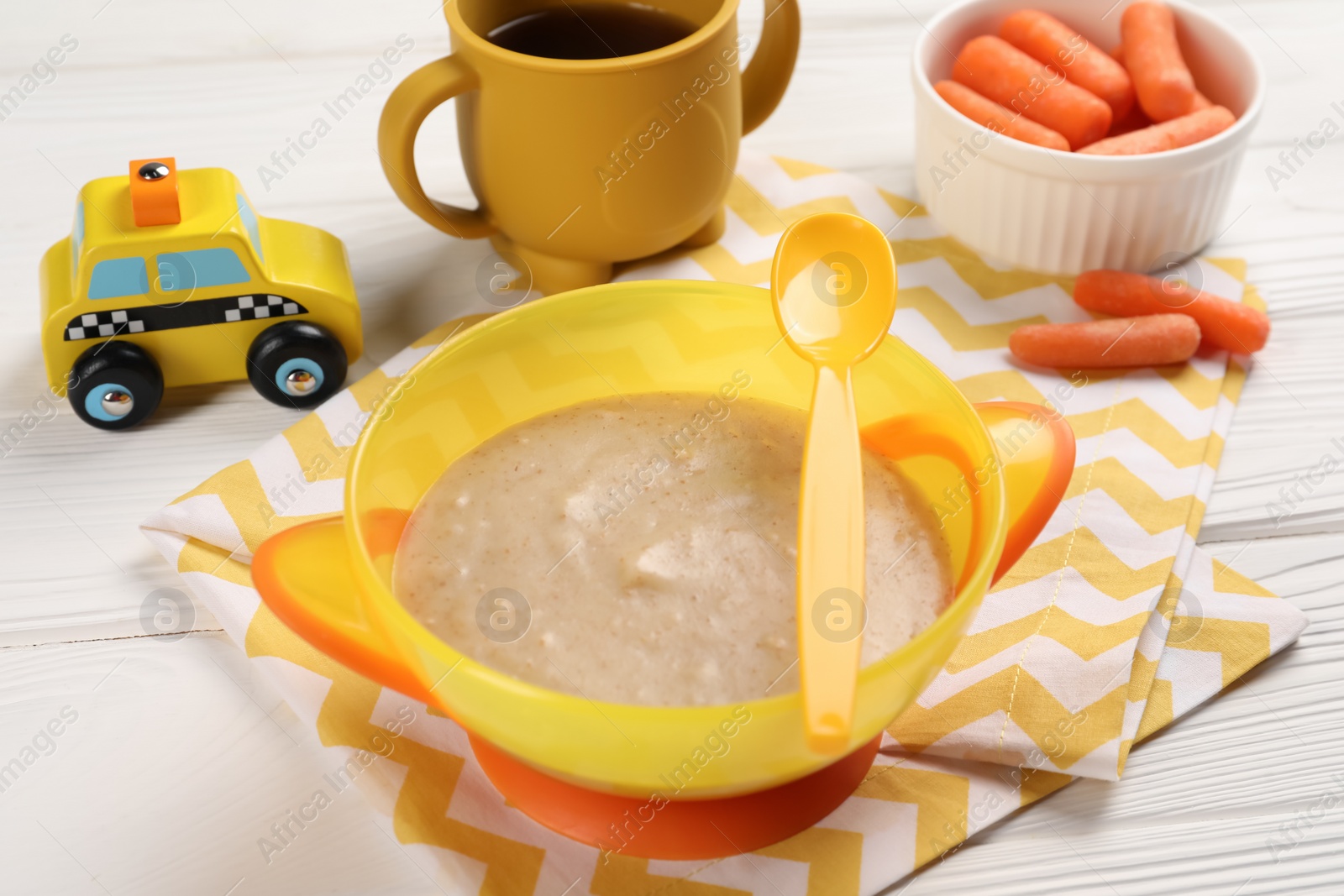 Photo of Baby food. Puree in bowl, small carrots, toy and drink on white wooden table