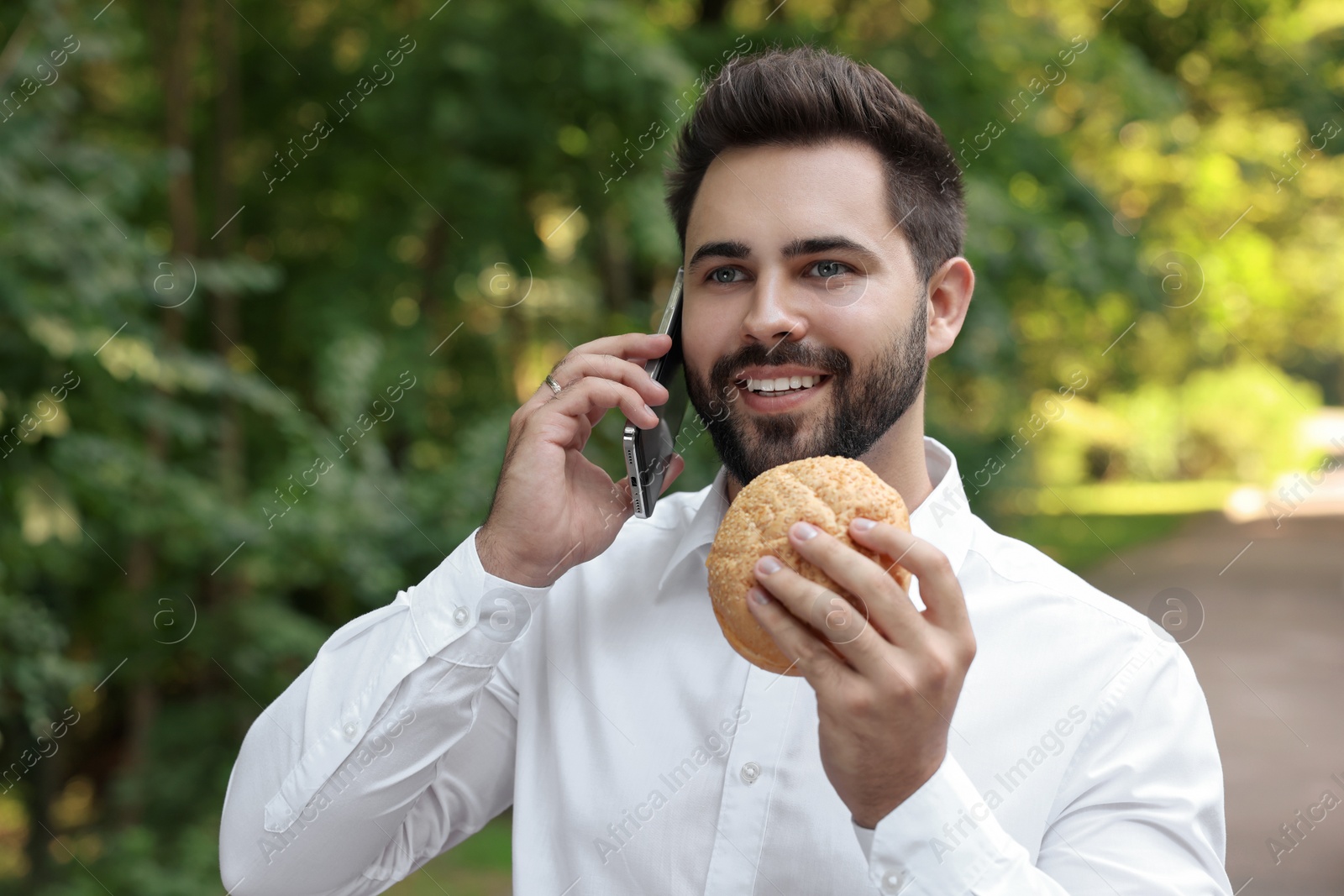 Photo of Lunch time. Young businessman with hamburger talking on smartphone in park