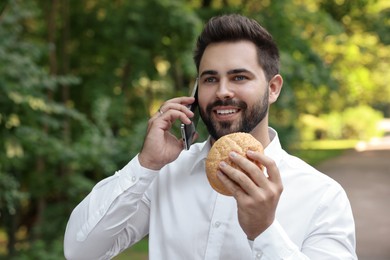 Photo of Lunch time. Young businessman with hamburger talking on smartphone in park