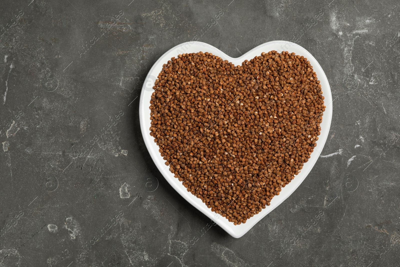 Photo of Heart shaped plate with raw buckwheat on grey background, top view