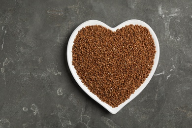 Heart shaped plate with raw buckwheat on grey background, top view