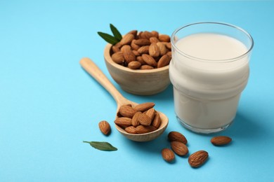 Fresh almond milk in glass, nuts, spoon and green leaves on light blue background, space for text