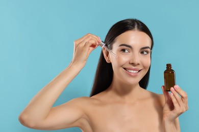 Beautiful young woman applying serum onto her face on light blue background