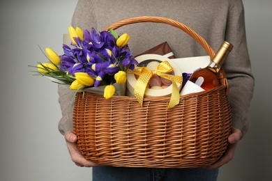 Photo of Man holding wicker basket with gifts, bouquet and wine on grey background, closeup