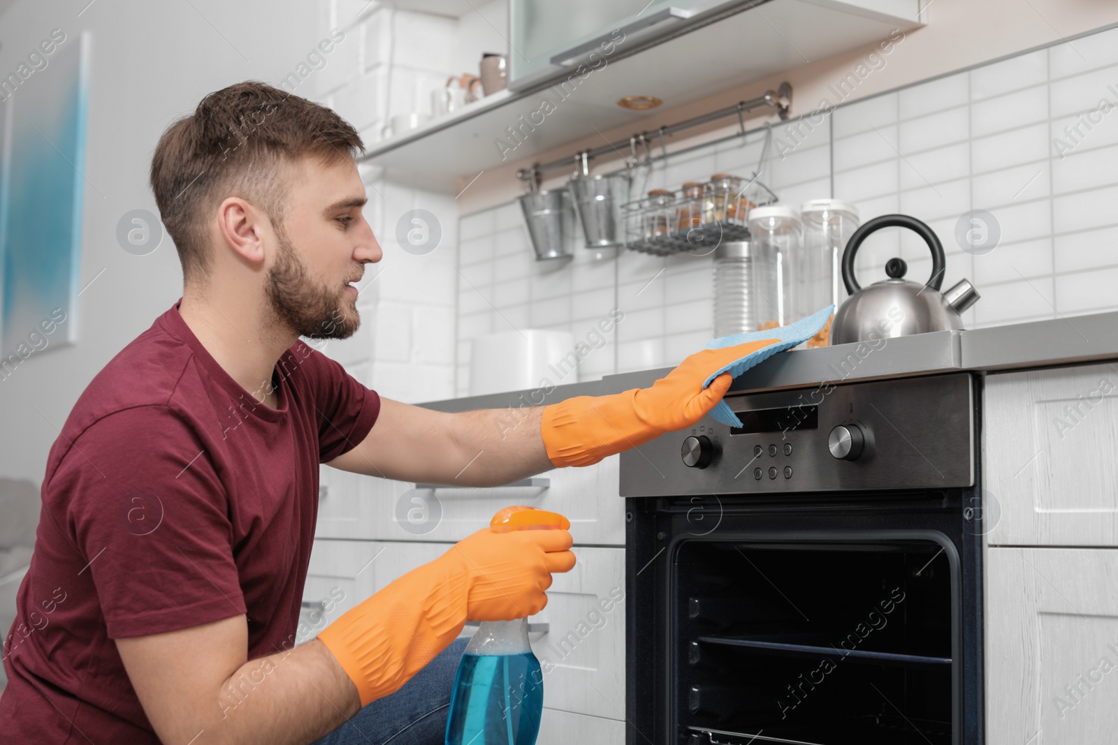 Photo of Young man cleaning oven with rag and detergent in kitchen