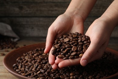 Photo of Woman taking pile of roasted coffee beans from bowl at wooden table, closeup