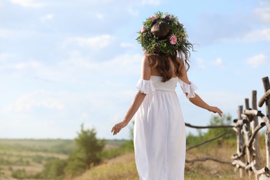 Young woman wearing wreath made of beautiful flowers outdoors on sunny day, back view