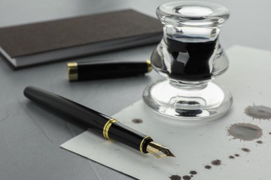 Photo of Stylish fountain pen, paper with blots of ink and inkwell on light grey textured table