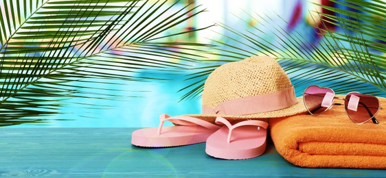 Beach towel, flip flops, straw hat and heart shaped sunglasses on light blue wooden surface near outdoor swimming pool. Banner design