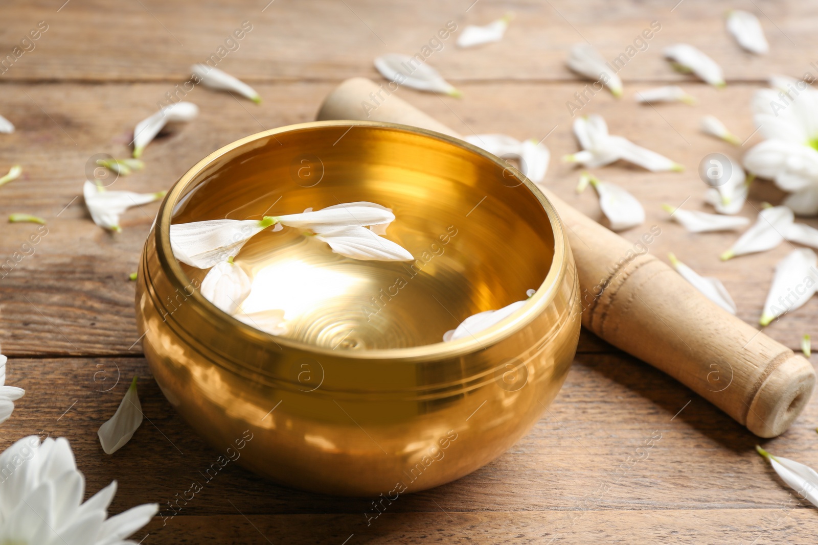 Photo of Golden singing bowl with petals and mallet on wooden table, closeup. Sound healing