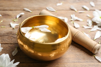 Golden singing bowl with petals and mallet on wooden table, closeup. Sound healing