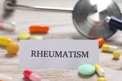 Card with word Rheumatism, pills and stethoscope on white wooden table, closeup