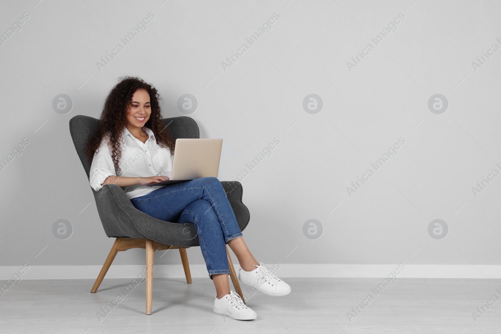 Photo of Young African-American woman working on laptop in armchair indoors. Space for text
