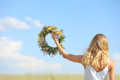 Photo of Young woman holding wreath made of flowers in field on sunny day, back view