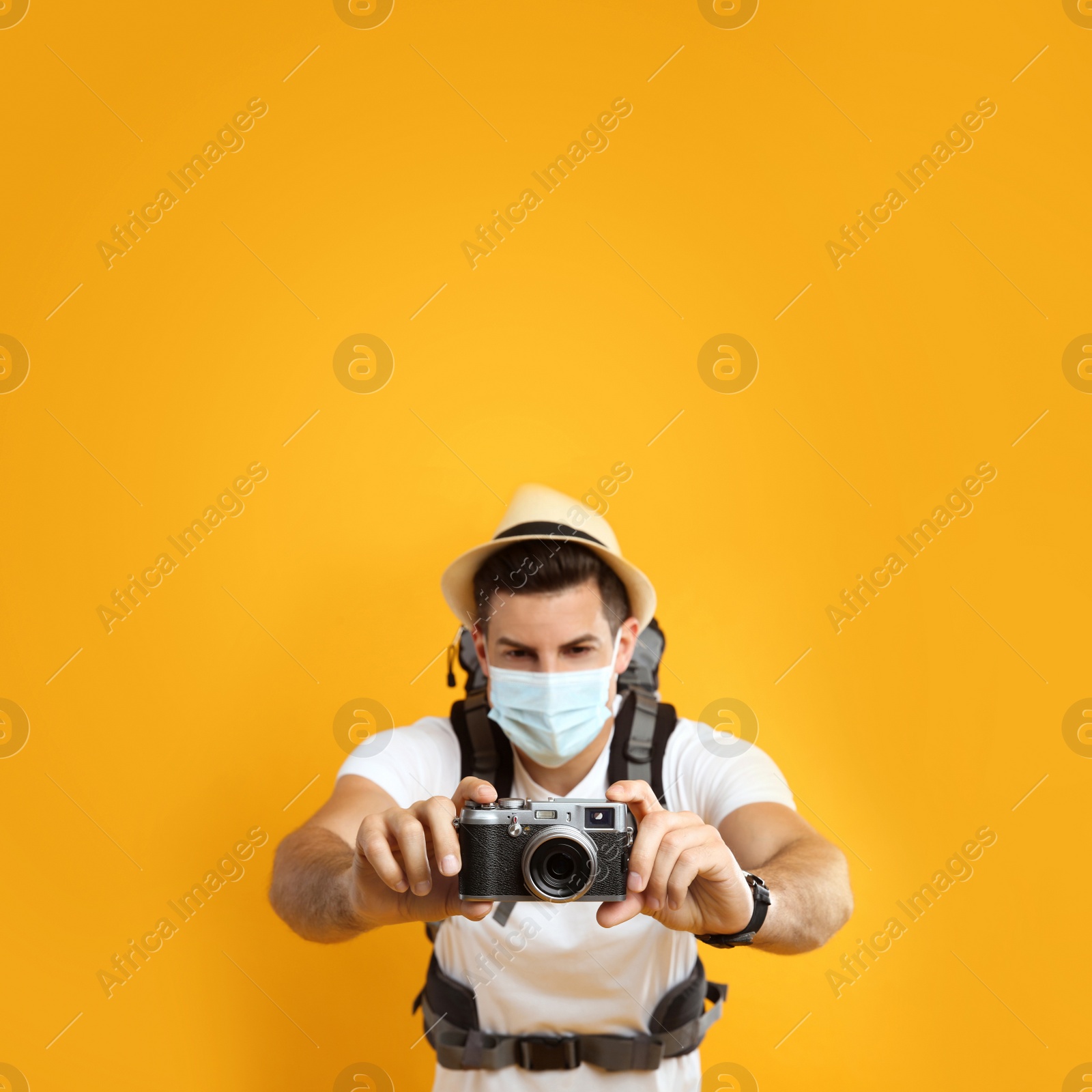 Photo of Male tourist in protective mask with travel backpack taking picture against yellow background, focus on camera