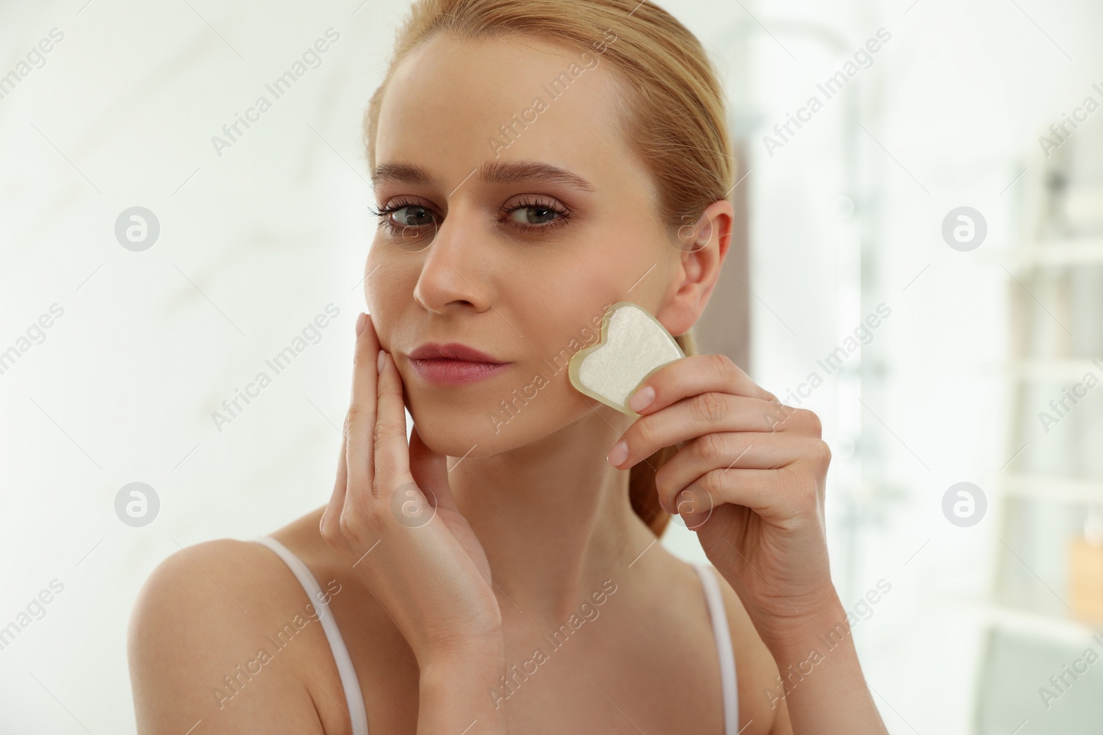 Photo of Beautiful young woman doing facial massage with gua sha tool in bathroom, closeup