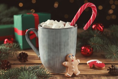 Photo of Delicious hot chocolate with marshmallows, candy cane and Christmas cookie on wooden table against blurred lights