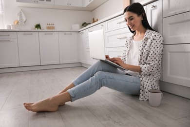 Photo of Happy woman with laptop and cup of drink sitting on warm floor in kitchen. Heating system